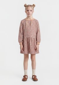 Ragazza giovane con doppio chignon biondo e trecce, indossa un vestito rosa floreale a maniche lunghe, calzini crema e scarpe marroni con cinturino, in piedi davanti a uno sfondo bianco.