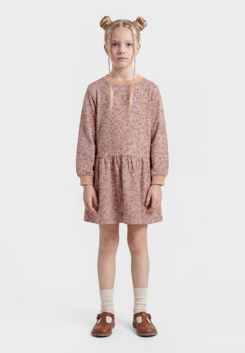 Ragazza giovane con doppio chignon biondo e trecce, indossa un vestito rosa floreale a maniche lunghe, calzini crema e scarpe marroni con cinturino, in piedi davanti a uno sfondo bianco.