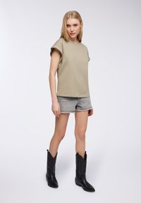 Top khaki a maniche corte abbinato a pantaloni corti in denim grigi strappati e stivali da cowboy neri. Gli stivali presentano cuciture decorative e una punta affusolata.