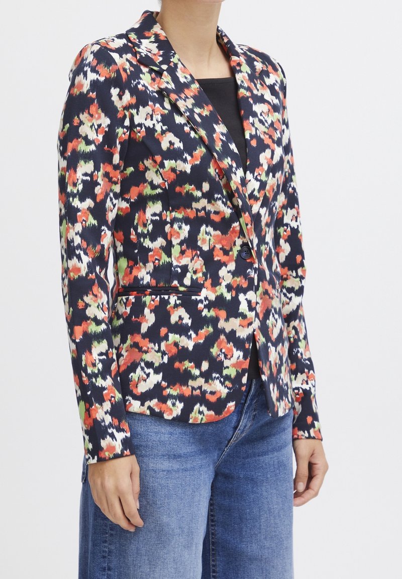 Femme portant un blazer ajusté à imprimé floral abstrait bleu marine, orange, vert et blanc, avec un jean bleu et un haut noir.