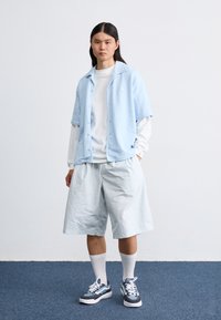 Camisa de botão de manga curta azul claro, blusa de manga longa branca por baixo, calções azuis claros a condizer e ténis escuros com detalhes brancos.