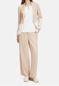 Blazer beige con maniche a contrasto crema, abbinato a una maglietta bianca a coste e pantaloni beige larghi. Il dettaglio distintivo è il bottone singolo del blazer.