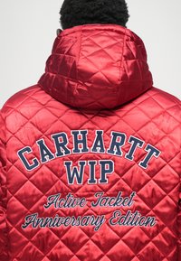Κόκκινο καπιτονέ μπουφάν με κουκούλα, διακοσμημένο με ναυτικό κεντημένο κείμενο "CARHARTT WIP" και "Active Jacket Anniversary Edition" �στην πλάτη.