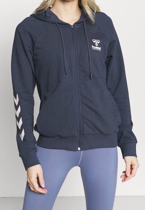 Haut à capuche zippé marine avec fermeture éclair à l'avant, capuchon réglable avec cordon de serrage et motif en chevron argenté sur la manche gauche. Texture du tissu douce.