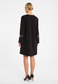 Schwarzes Kleid mit lockerer Passform, langen Ärmeln, die schwarze Lederakzente an den Bündchen aufweisen, und einem Rundhalsausschnitt. Getragen mit schwarzen High Heels.