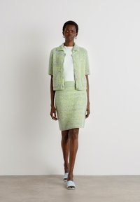 Vihreä ja sininen kuviollinen tweed-takki, jossa on lyhyet hihat, yhdistettynä samanlaiseen napakasti istuvaan hameeseen ja vaaleansinisiin liukumäkiin. Sileä pinta, napin yksityiskohdat.