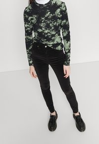 Czarna i zielona bluzka tie-dye z długim rękawem i prążkowaną fakturą, w zestawie z czarnymi wąskimi spodniami z aksamitu i czarnymi butami wsuwanymi z frędzlami.