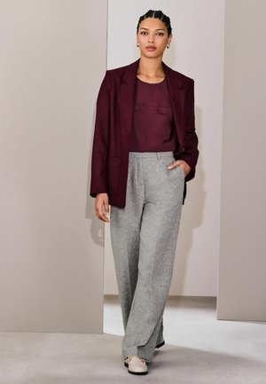 Punapäine patsidega naine kannab burgundia tooni bleiserit ja toppi, mida täiendavad kõrge vöökohaga hallid laia säärega püksid ja heledavärvilised kingad, seistes tavalise seinaga taustal.