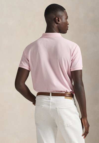 Άνδρας που κοιτάζει προς τα πίσω, φοράει ροζ κοντομάνικο πόλο πουκάμισο και άσπρο τζιν με καφέ ζώνη, πάνω σε απλό φόντο.