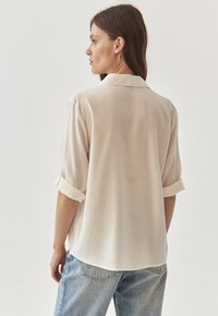 Light beige silk shirt with a soft texture, short rolled sleeves, and a collared neckline. The back has a curved hem and a relaxed fit.