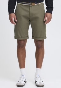 Shorts en coton vert olive avec ourlets retournés, dotés d'une ceinture avec une boucle en métal ronde et de détails rayés. Portés avec des chaussettes blanches et des baskets.