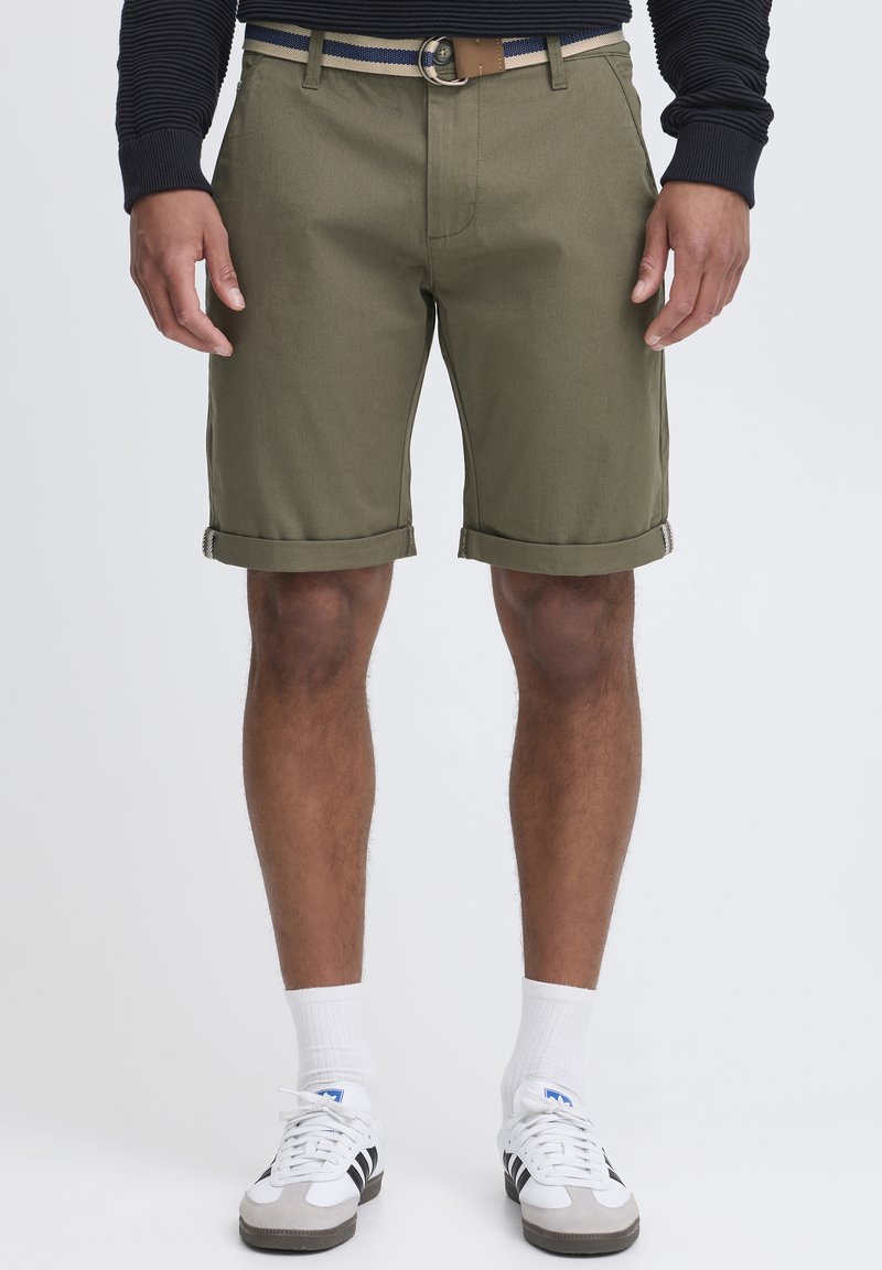 Shorts en coton vert olive avec ourlets retournés, dotés d'une ceinture avec une boucle en métal ronde et de détails rayés. Portés avec des chaussettes blanches et des baskets.