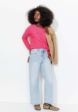Pull en tricot rose vif, surchemise beige, jean large bleu clair et chaussures décontractées à imprimé léopard. Le mannequin pose devant un fond blanc.