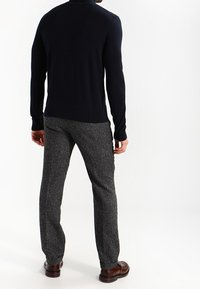 Pull tricoté bleu marine à col montant, associé à un pantalon gris texturé. La tenue présente une silhouette ajustée et des chaussures marron.