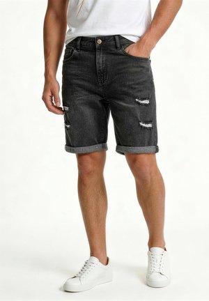 Mann trägt hochgekrempelte schwarze, abgenutzte Jeansshorts, weiße Sneakers und ein weißes T-Shirt, steht mit einer Hand in der Tasche.