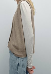 Beige neuleliivi avoimella edessä, tekstuuri-kankaasta, käytetty pitkähihainen vaaleanpainen päällä. Yhdistetty vaaleansinihin farkkuihin.