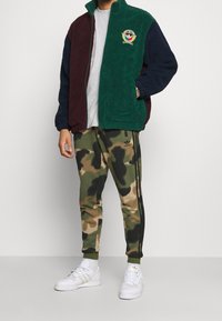 Fleecová bunda v zelené, vínové a námořnické modři s vyšívaným logem; maskáčové tepláky s černými bočními pruhy; bílé tenisky se zlatými detaily.