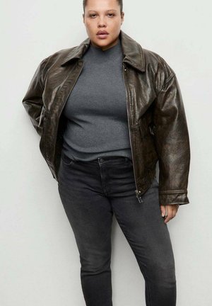 Veste en cuir marron à finition brillante, grand col et fermeture éclair, portée par-dessus une chemise grise et un jean noir. Texture lisse et coupe décontractée.