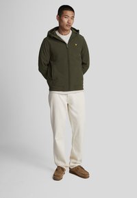 Olivenfarget zip-jakke med hette, ribbede mansjetter og hem, med en liten brodert logo på brystet. Kombinert med kremfargede bukser.