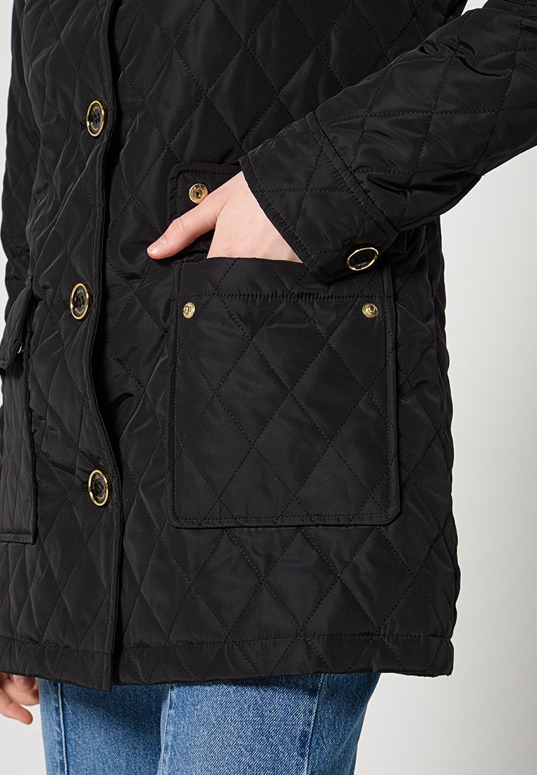 Veste matelassée noire avec un motif en losange, du matériel doré et une poche à rabat, avec une main placée dans la poche latérale.