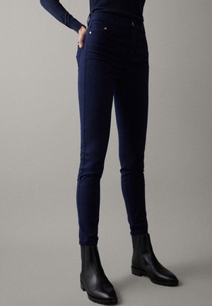 Person, die dunkelblaue, hoch taillierte Skinny-Hosen mit schwarzen Stiefeletten trägt, vor einem neutralen, einfarbigen Hintergrund stehend.