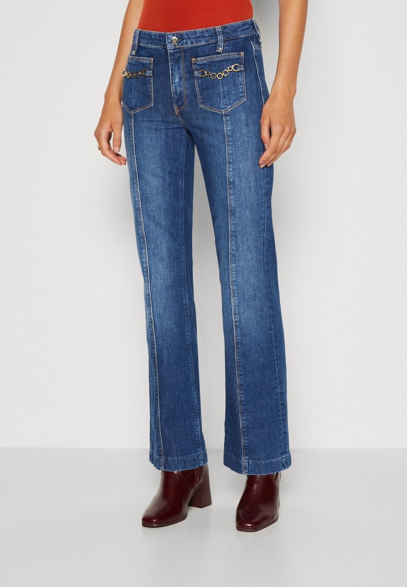 Femme portant un jean bleu droit avec un détail chaîne dorée sur les poches avant et des bottines marron à talons, debout devant un fond uni.