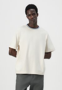 Beige kortermet t-skjorte med rund hals og blå og hvite striper som dekorasjon. Mykt stoff med en avslappet passform og rett kant.