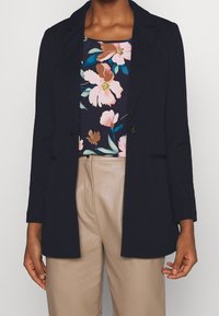 Blazer bleu marine à manches longues, fermeture à un bouton et revers. Porté sur un haut à motif floral avec des accents roses et bleus. Texture lisse.