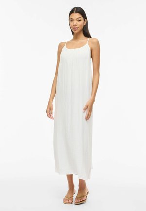 Kobieta stojąca w białej, bezrękawnikowej sukience do kostek z cienkimi ramiączkami i jasno-brązowych, odkrytych sandałach na białym tle.