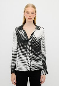 Blusa a righe bianco e nero con maniche lunghe, colletto e chiusura a bottone. Presenta un design a pois sfumati con una texture liscia.