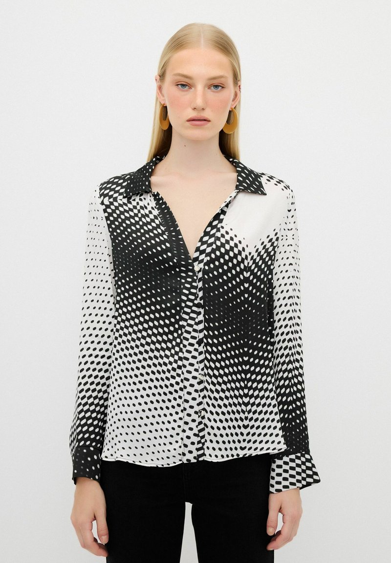 Blusa a righe bianco e nero con maniche lunghe, colletto e chiusura a bottone. Presenta un design a pois sfumati con una texture liscia.