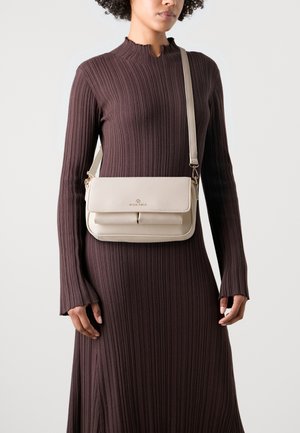 Bolso bandolera beige con solapa y herrajes dorados, que cuenta con dos bolsillos frontales y una textura suave. Llevado sobre un vestido ajustado marrón de canalé.
