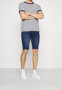 Hombre de pie con los brazos cruzados, vistiendo una camiseta a rayas blancas y marinas, pantalones cortos de mezclilla azul oscuro, calcetines blancos y zapatillas blancas.