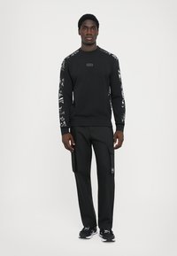 Sweatshirt preto com mangas padrões, decote redondo e um emblema. Combinado com calças cargo pretas e ténis pretos.