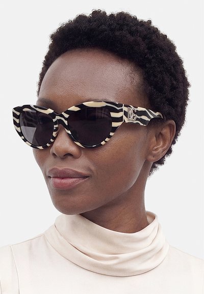 Frau mit kurzen lockigen Haaren, die schwarz-weiße, zebragestreifte Katzenaugen-Sonnenbrille und ein helles Top mit hohem Kragen trägt.