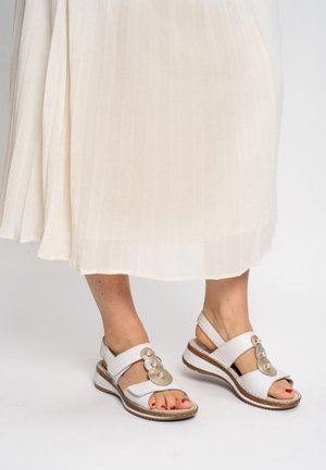 Weiße Ledersandalen mit geschlossenem Fersenbereich, dekorativen runden Akzenten und einer strukturierten Sohle. Kombiniert mit einem plissierten cremefarbenen Rock.