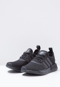 Schwarze Sportschuhe mit einem Netzoberteil, drei gestreiften Akzenten, runder Zehenpartie, strukturiertem Sohlenprofil und Zuglaschen an der Ferse und der Zunge.