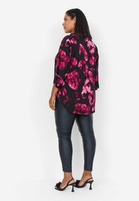 Haut noir avec un grand motif floral rose, coupe ample, manches trois-quarts, associé à des leggings noirs brillants et des sandales à talons noires.