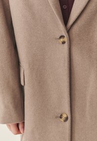Beiger Mantel aus Wollmischung mit Reverskragen, zwei Knöpfen und weicher Textur. Verfügt über Seitentaschen und eine strukturierte Passform.