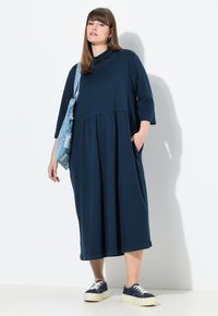 Tummansininen, polvipituinen mekko korkealla kauluksella, 3/4-hihalla, kootulla vyötäröllä ja taskuilla. Yhdistetty sinisiin tennareihin ja farkkusalin laukkuun.