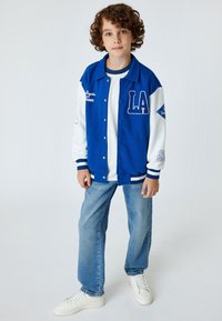 Giacca varsity blu con maniche bianche, con la scritta "LA", abbinata a una maglietta bianca e jeans azzurri chiari. Sneakers bianche completano l'outfit.