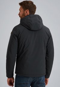 Chaqueta negra con capucha y un acabado suave, que presenta un cuello alto y detalles cosidos. La parte trasera muestra un diseño sin costuras y un ajuste relajado.