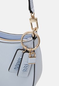 Lyseblå håndtaske med tekstureret materiale, guldfarvet hardware og en nøglering. Har dekorative mærker med teksten "GUESS" og "1981".