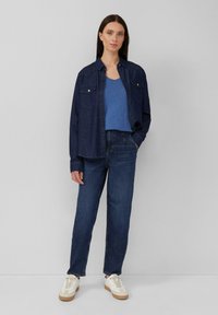 Camicia di jeans blu scuro con due tasche sul petto, indossata su una canotta blu aderente. Jeans blu scuro larghi e sneakers bianche con suola in gomma.