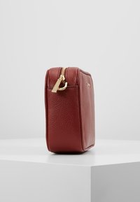 Mała prostokątna torebka z teksturowanej ciemnoczerwonej skóry z złotym zamkiem błyskawicznym i detalem logo, stojąca pionowo na białej powierzchni.