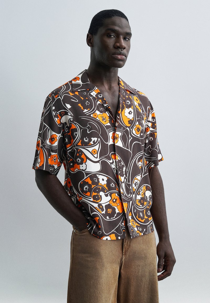 Άνδρας που φοράει κοντομάνικο πουκάμισο με κουμπιά, με καφέ, πορτοκαλί και λευκό αφηρημένο λουλουδένιο μοτίβο, και μπεζ παντελόνι, στέκεται μπροστά σε μια απλή φόντο.