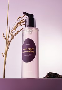 Olej oczyszczający w przezroczystej butelce z czarną pompką. Etykieta w ciemnym fioletowym kolorze ze złotym tekstem. Towarzyszą mu ziarna czarnego ryżu.