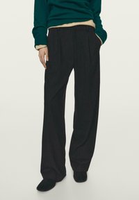 Schwarze Nadelstreifenhose mit weitem Schnitt, vorne mit Falten und seitlichen Taschen, kombiniert mit einem dunkelgrünen Pullover.