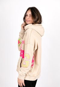 Beige hoodie featuring colorful star and number graphics, with a textured finish and a hood. The design includes accents in pink and green.