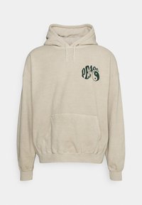 Sudadera beige con bolsillo frontal, capucha y ajuste holgado. Presenta un texto gráfico en verde oscuro que dice "PAZ" con un diseño de yin-yang en el lado izquierdo del pecho.